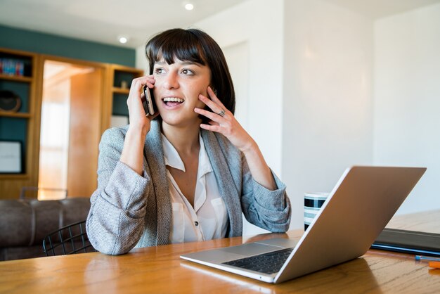 Ritratto di giovane donna che parla sul suo telefono cellulare e lavora da casa con il computer portatile. Concetto di ufficio a casa. Nuovo stile di vita normale.