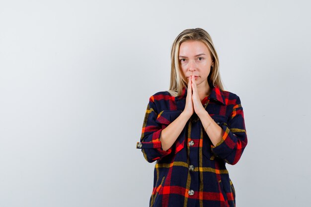 Ritratto di giovane donna che preme le mani per pregare in camicia controllata e guardando la vista frontale speranzoso