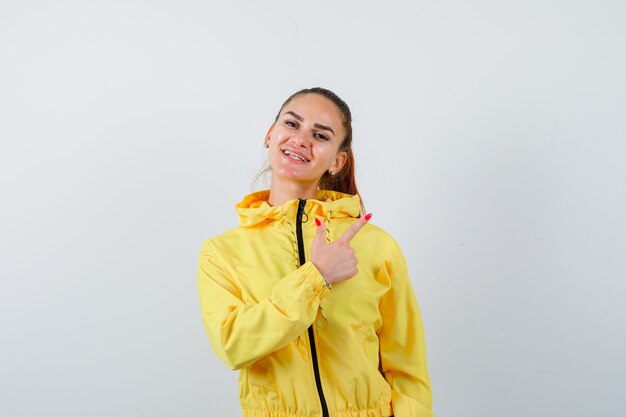 Ritratto di giovane donna che punta all'angolo in alto a destra in giacca gialla e guarda una vista frontale allegra