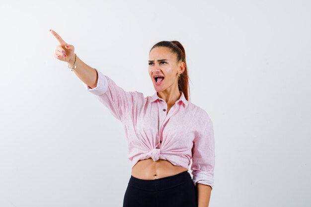 Ritratto di giovane donna che punta lontano in camicia, pantaloni e guardando eccitato vista frontale