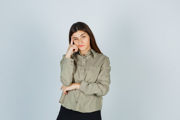 Ritratto di giovane donna che si appoggia con la testa a portata di mano, incurvando le labbra in camicia, gonna e guardando una vista frontale pensierosa