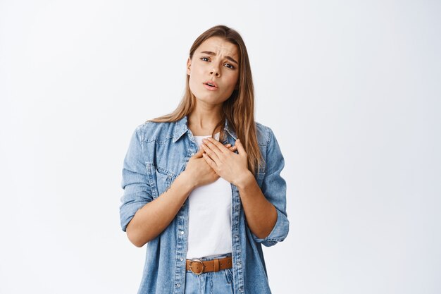 Ritratto di giovane donna che si tiene per mano sul cuore e guarda qualcosa di adorabile e bello, sentendosi commossa e sincera, in piedi sul muro bianco