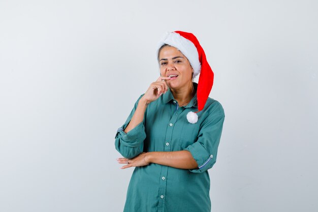 Ritratto di giovane donna che tiene il dito sul labbro inferiore in camicia, cappello da Babbo Natale e guarda una vista frontale pensierosa