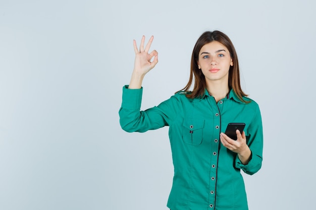 Ritratto di giovane donna che tiene il telefono cellulare, mostrando il gesto giusto in camicia verde e guardando la vista frontale soddisfatta