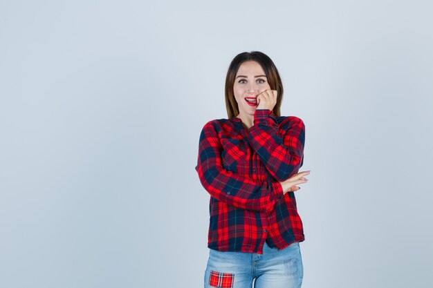 Ritratto di giovane donna che tiene la mano sulla guancia in camicia a quadri, jeans e guarda stupita vista frontale