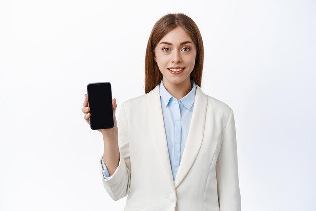 Ritratto di giovane donna d'affari mostra schermo mobile vuoto e sorrisi ha dimostrato che l'applicazione finanziaria si trova su sfondo bianco