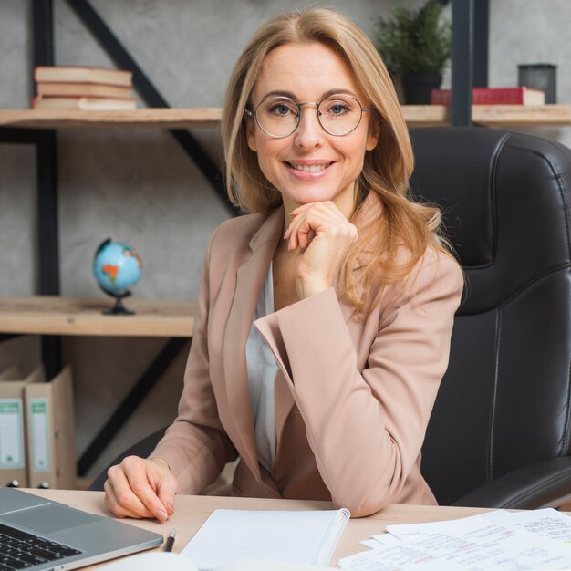 Ritratto di giovane donna di affari bionda sicura che si siede sulla sedia nel luogo di lavoro