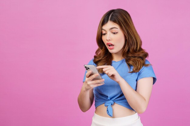 Ritratto di giovane donna eccitata che indossa una maglietta casual utilizzando lo smartphone isolato su sfondo rosa