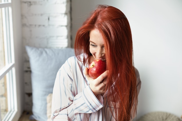 Ritratto di giovane donna europea affascinante e gioiosa rossa che indossa pigiama a righe alla moda con sguardo felice, morde mela rossa matura fresca, mangia frutta per colazione, seduto vicino alla finestra nella sua stanza