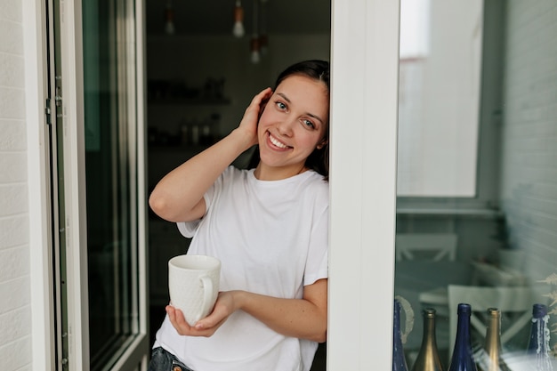 Ritratto di giovane donna europea con capelli scuri e pelle sana sorridente con il caffè del mattino nella moderna cucina leggera.