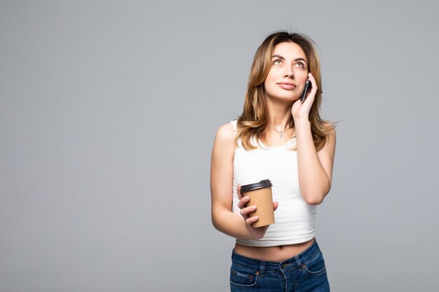 Ritratto di giovane donna felice che posa conversazione dal caffè bevente del telefono cellulare isolato sopra la parete grigia