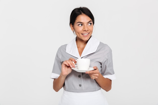 Ritratto di giovane donna felice del brunette in uniforme grigia che tiene tazza di tè e che osserva da parte