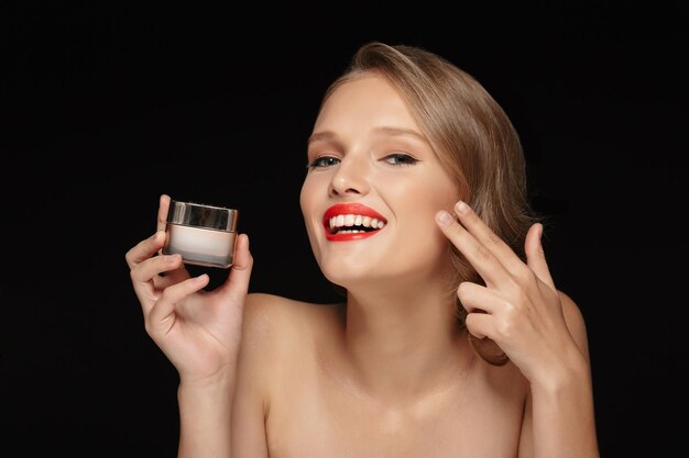 Ritratto di giovane donna gioiosa con capelli mossi e labbra rosse che tiene in mano una crema di bellezza mentre guarda felicemente in camera su sfondo nero