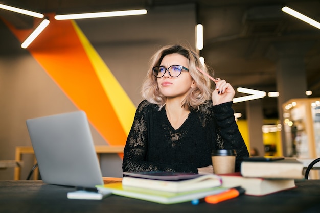 Ritratto di giovane donna graziosa che si siede al tavolo in camicia nera che lavora al computer portatile in ufficio di co-working, con gli occhiali, pensando al problema