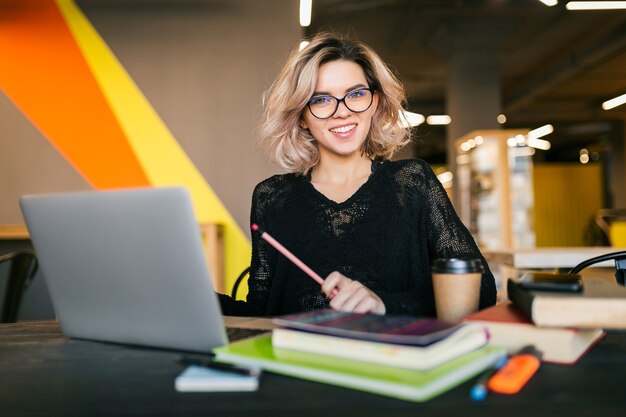 Ritratto di giovane donna graziosa che si siede al tavolo in camicia nera che lavora al computer portatile in ufficio di co-working