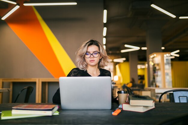 Ritratto di giovane donna graziosa che si siede al tavolo in camicia nera che lavora al computer portatile in ufficio di co-working