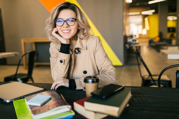 Ritratto di giovane donna graziosa che si siede al tavolo in trench lavorando sul portatile in ufficio di co-working, con gli occhiali, sorridente, felice, positivo, sul posto di lavoro