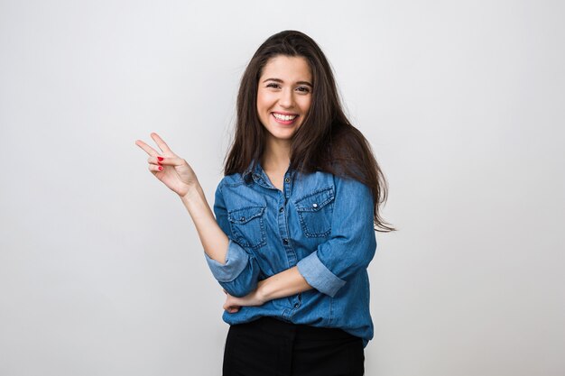 Ritratto di giovane donna graziosa che sorride in camicia di jeans blu, umore felice e positivo, sorriso isolato e sincero, capelli lunghi, che mostra il segno di pace, umore positivo