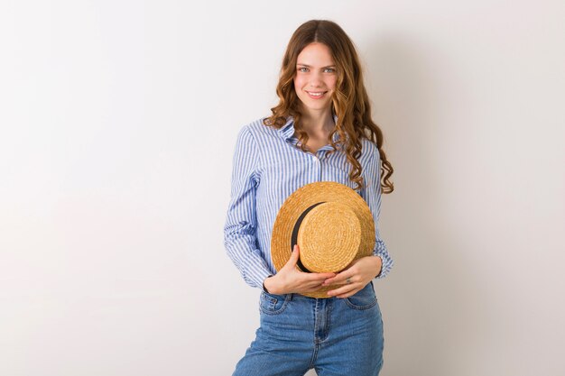 Ritratto di giovane donna graziosa con camicia di cotone blu jeans cappello di paglia in posa sul muro bianco