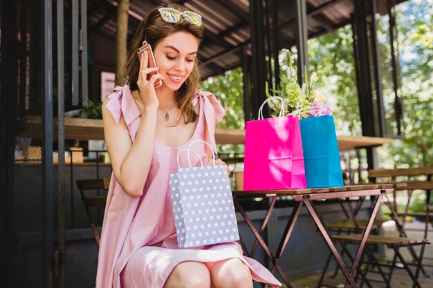 Ritratto di giovane donna graziosa felice sorridente con espressione del viso sorpreso seduto nella caffetteria con borse della spesa parlando al telefono, vestito di moda estiva, abito di cotone rosa, abbigliamento alla moda