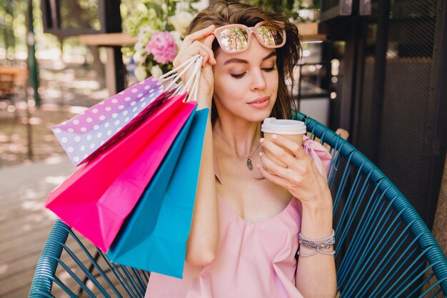 Ritratto di giovane donna graziosa felice sorridente con espressione faccia eccitata seduto nella caffetteria con borse della spesa bere caffè, vestito di moda estiva, abito di cotone rosa, abbigliamento alla moda