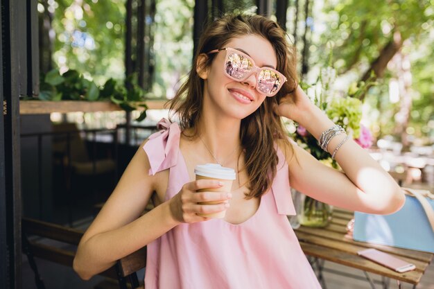 Ritratto di giovane donna graziosa felice sorridente con seduta in caffè bere caffè, abito di moda estiva, abito di cotone rosa, accessori di abbigliamento alla moda