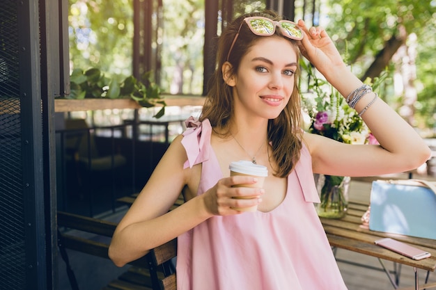 Ritratto di giovane donna graziosa felice sorridente con seduta in caffè bere caffè, vestito di moda estiva, stile hipster, vestito di cotone rosa, accessori di abbigliamento alla moda