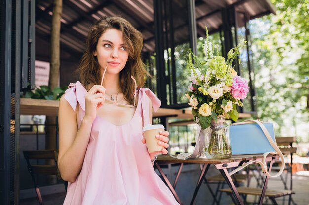 Ritratto di giovane donna graziosa felice sorridente con seduta in caffè bere caffè, vestito di moda estiva, stile hipster, vestito di cotone rosa, accessori di abbigliamento alla moda