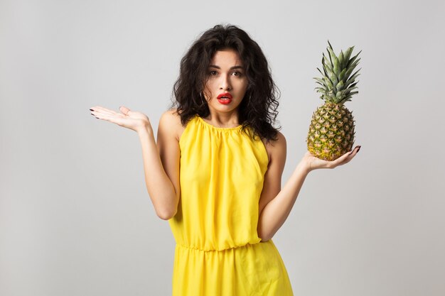 Ritratto di giovane donna graziosa sorpresa in vestito giallo, che tiene ananas, emozione divertente, espressione del viso scioccato, stile estivo, dieta di frutta, guardando a porte chiuse, razza mista, isolato, alzando le mani