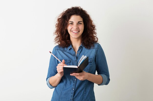 Ritratto di giovane donna graziosa sorridente hipster naturale con l'acconciatura riccia in camicia di jeans in posa con il taccuino e la penna isolato su sfondo bianco studio, apprendimento degli studenti