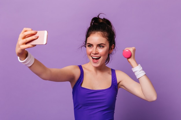 Ritratto di giovane donna in fitness top tenendo il manubrio rosa e tenendo selfie sulla parete viola