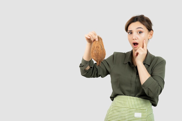 Ritratto di giovane donna in grembiule che tiene una noce di cocco contro il muro bianco