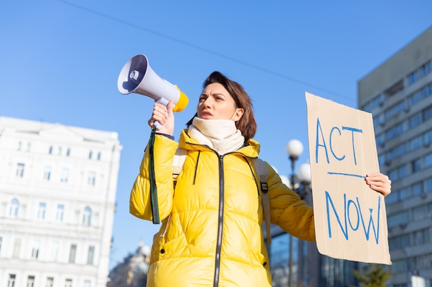 Ritratto di giovane donna in piedi all'aperto in città e mostrando la tabella Act Now. Scheda di dimostrazione femminile con protesta contro questioni pandemiche, politiche o ambientali. Protesta unica.