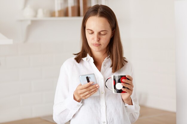 Ritratto di giovane donna infelice adulta in piedi con lo smartphone in mano, bevendo caffè o tè in cucina, esprimendo emozioni negative durante la lettura del messaggio.