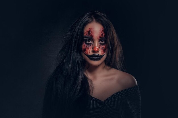 Ritratto di giovane donna nel ruolo di malvagio clown spaventoso in studio fotografico scuro.