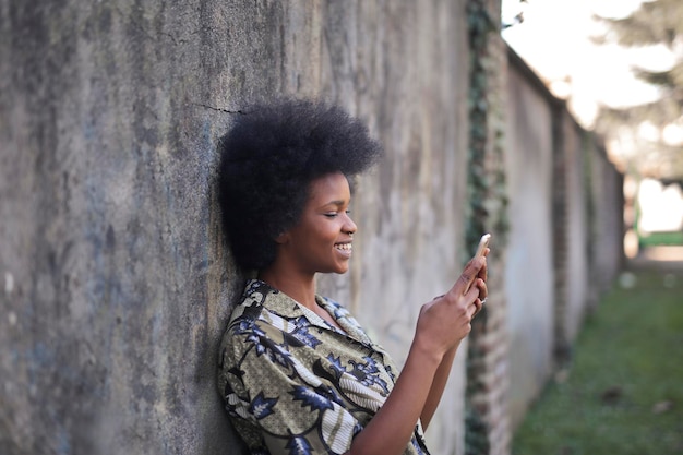 ritratto di giovane donna nera appoggiata a un muro utilizzando il suo smartphone