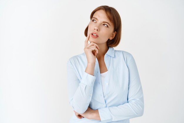 Ritratto di giovane donna premurosa che guarda da parte il logo pensando e facendo la scelta ponderando la decisione in piedi in camicetta su sfondo bianco