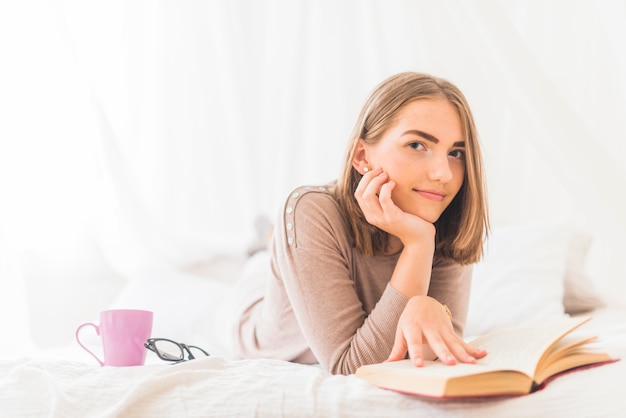 Ritratto di giovane donna sdraiata sul letto per leggere il libro con il caffè