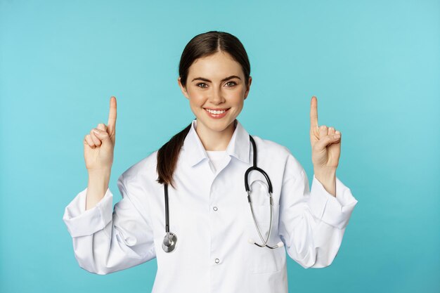 Ritratto di giovane donna sicura di sé medico, operatore medico in cappotto, che punta il dito verso l'alto e sorridente, mostrando pubblicità di medicinali, promozione clinica, sfondo toquoise