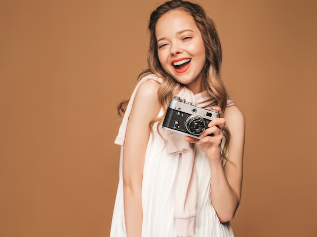 Ritratto di giovane donna sorridente allegra che prende foto con ispirazione e che porta vestito bianco. Ragazza che tiene la retro macchina fotografica. Posa di modello