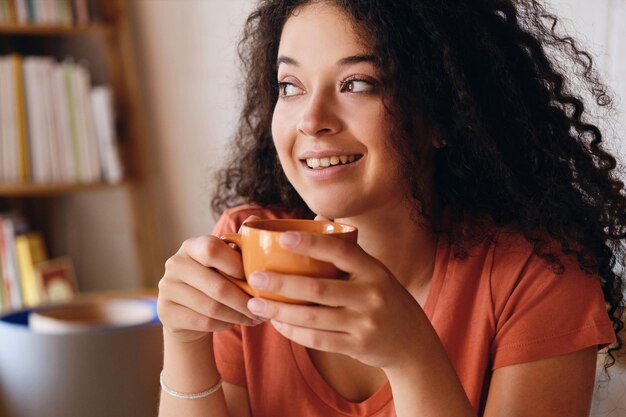 Ritratto di giovane donna sorridente attraente con capelli ricci scuri che tiene in mano una tazza di caffè arancione che guarda felicemente da parte nella casa moderna e accogliente da solo