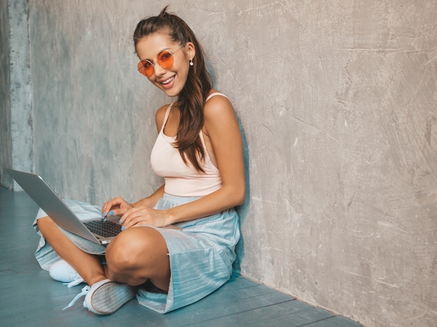 Ritratto di giovane donna sorridente creativa in occhiali da sole. Bella ragazza che si siede sul pavimento vicino alla parete grigia. Modello con notebook. Femmina vestita in abiti hipster