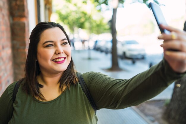 Ritratto di giovane donna taglie forti prendendo selfie con il suo telefono mophile all'aperto in strada. Concetto urbano.