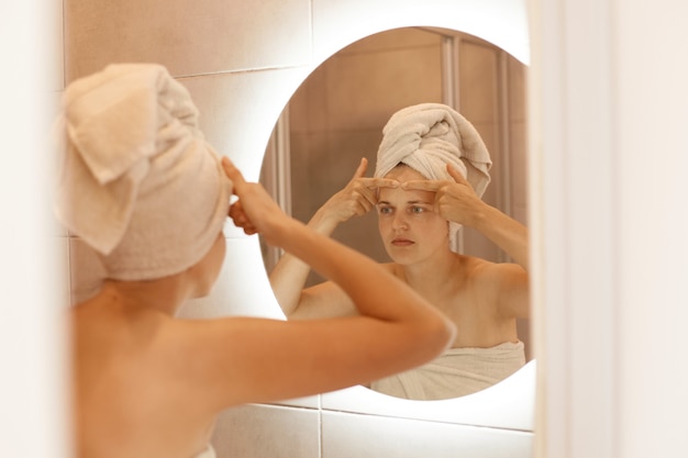 Ritratto di giovane donna turbata che controlla l'acne sulla fronte, guardando lo specchio del bagno con un'espressione facciale triste e preoccupata, essendo avvolta in un asciugamano bianco.