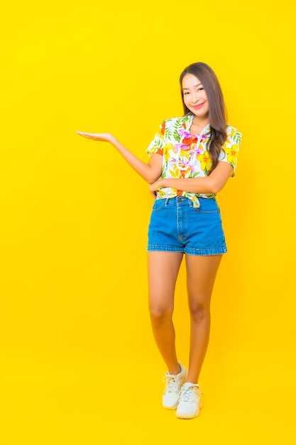 Ritratto di giovane e bella donna asiatica che indossa maglietta colorata e mostrando qualcosa sul muro giallo