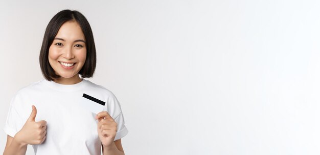Ritratto di giovane e bella donna asiatica moderna che mostra la carta di credito e i pollici in su consigliando il pagamento contactless in piedi su sfondo bianco
