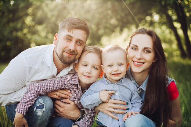 Ritratto di giovane famiglia felice seduto nel parco in ginocchio in posa e guardando la fotocamera con luci di sfondo del sole Madre e padre che abbracciano i loro due figli Concetto di famiglia amichevole