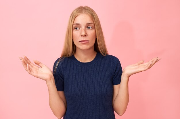 Ritratto di giovane femmina carina confusa perplessa essendo a corto di parole, scrollando le spalle e facendo un gesto della mano, avendo un'espressione facciale perplessa