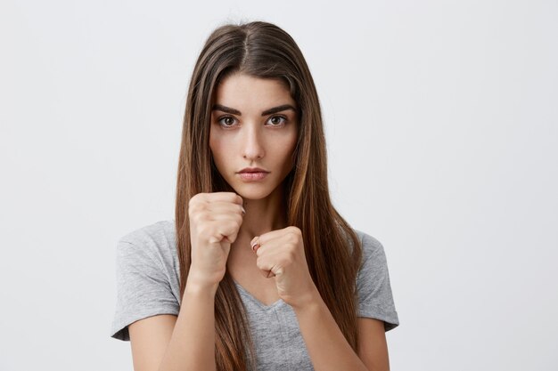 Ritratto di giovane femmina caucasica attraente seria con capelli lunghi scuri in maglietta grigia alla moda che tengono le mani combatto posa, con espressione media e concentrata