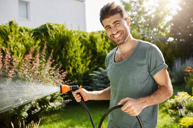 Ritratto di giovane fiorista gioioso attraente in maglietta blu sorridente con i denti, annaffiare i fiori in casa di campagna, facendo un lavoro rilassante in una mattina d'estate.